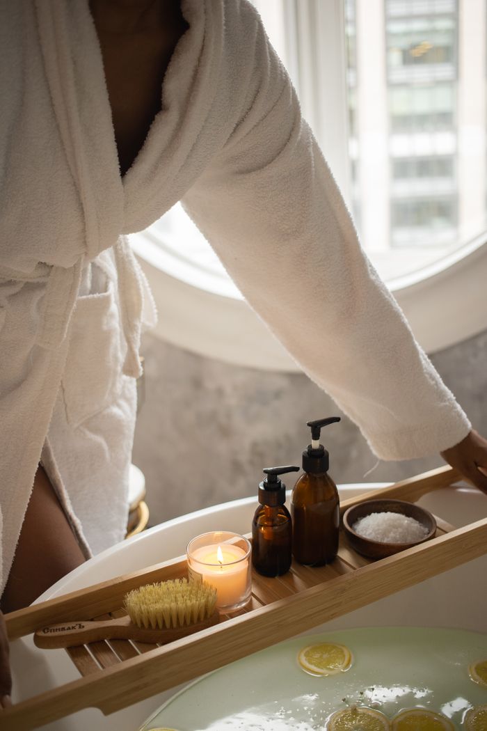 Faceless female in white bathrobe putting wooden tray with spa supplies and burning candle on tub while preparing for skincare procedure